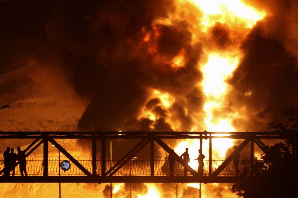 إيرانيون يراقبون من فوق جسر النيران المتصاعدة بعد هجوم إسرائيلي على مستودع نفط شاران(رويترز).