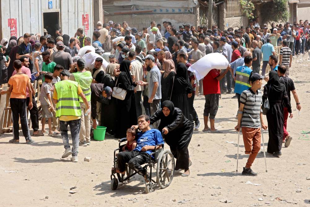 امرأة فلسطينية تدفع رجلاً وطفلاً على كرسي متحرك بينما يتجمع الناس لتلقي أكياس الدقيق في مستودع لبرنامج الأغذية العالمي في مدينة غزة (أ ف ب)