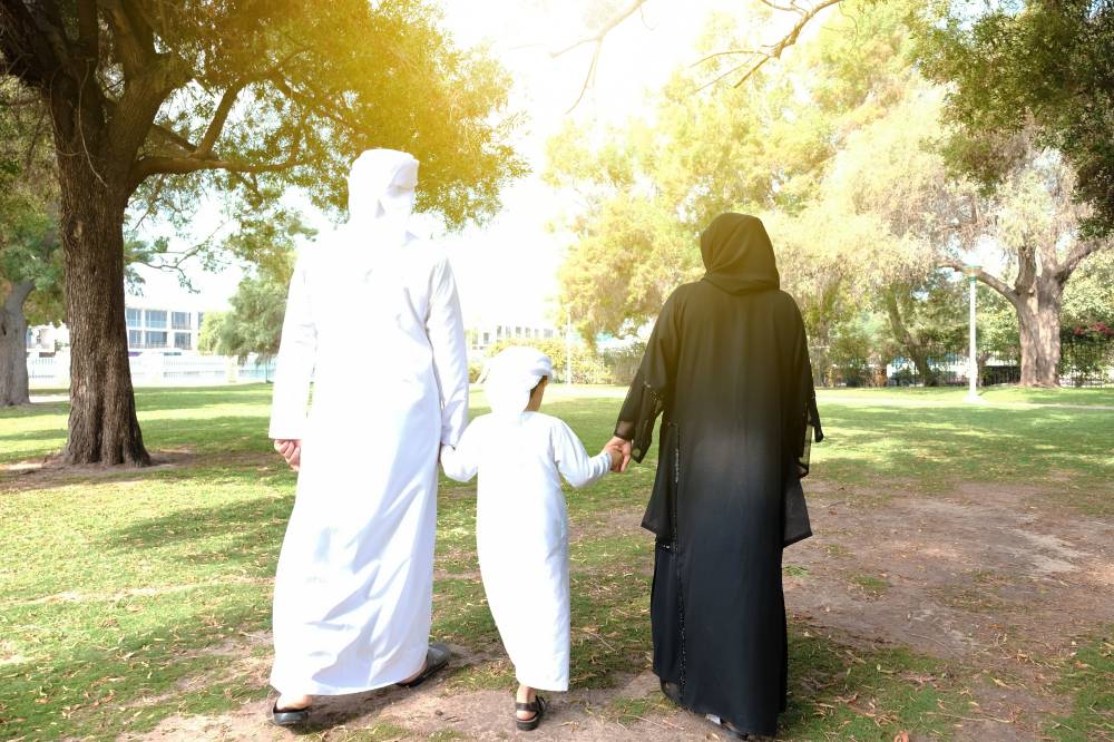 Back shot of Arab family for three. Rear view of Arabic man in dish dasha and Woman in Abaya with their son on Kandura. Walking together at a park with sunny day light