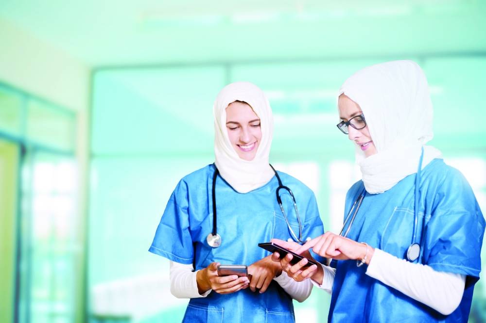 Two female middle eastern doctors in blue medical uniform standing in a hospital hallway, looking at their smart phones, smiling